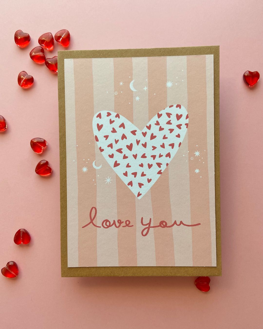 Carte postale illustrée au design romantique avec un grand cœur blanc parsemé de petits cœurs rouges sur un fond à rayures beige et rose. L'inscription "love you" en écriture manuscrite ajoute une touche délicate et élégante, idéale pour exprimer l'amour et la tendresse.