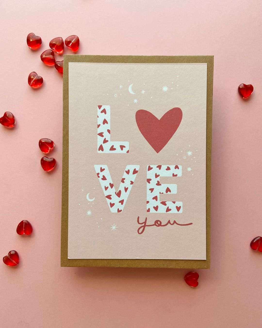 Carte postale illustrée au design romantique avec un grand cœur rouges sur un fond beige rosé. L'inscription "love you" en écriture manuscrite ajoute une touche délicate et élégante, idéale pour exprimer l'amour et la tendresse.