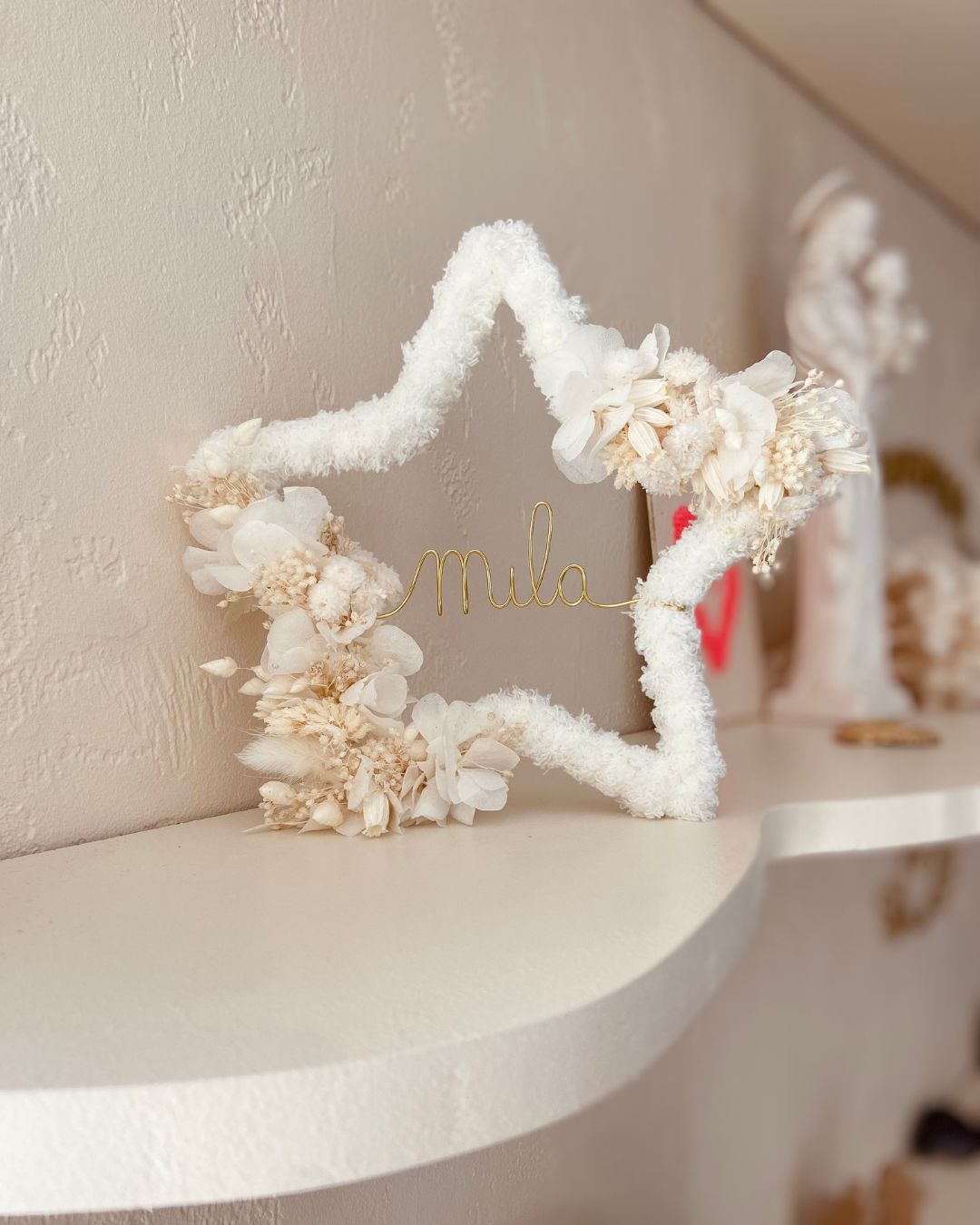 Présentation d'une étoile moumoute blanche, ornée de fleurs séchées blanches, modèle Ava, personnalisée, sur une étagère – une création élégante et intemporelle signée Mes Attrapes Rêves.