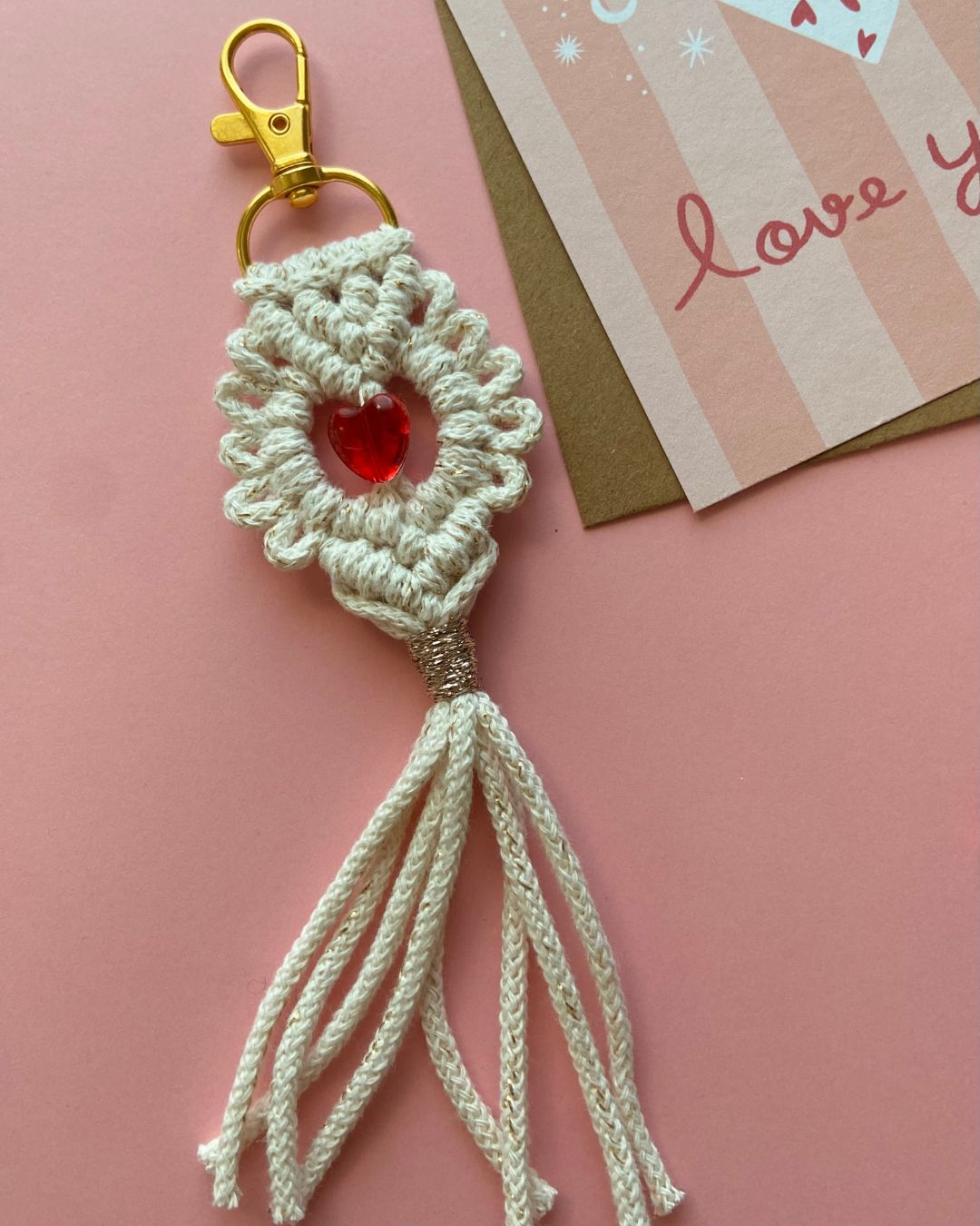 Porte-clés en macramé fait main, orné d’un cœur rouge en cristal et d’un mousqueton doré, sur fond rose avec des perles en forme de cœur dispersées.