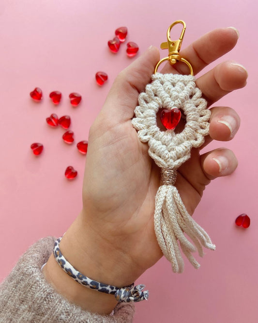 Porte-clés en macramé fait main, orné d’un cœur rouge en cristal et d’un mousqueton doré, sur fond rose avec des perles en forme de cœur dispersées.